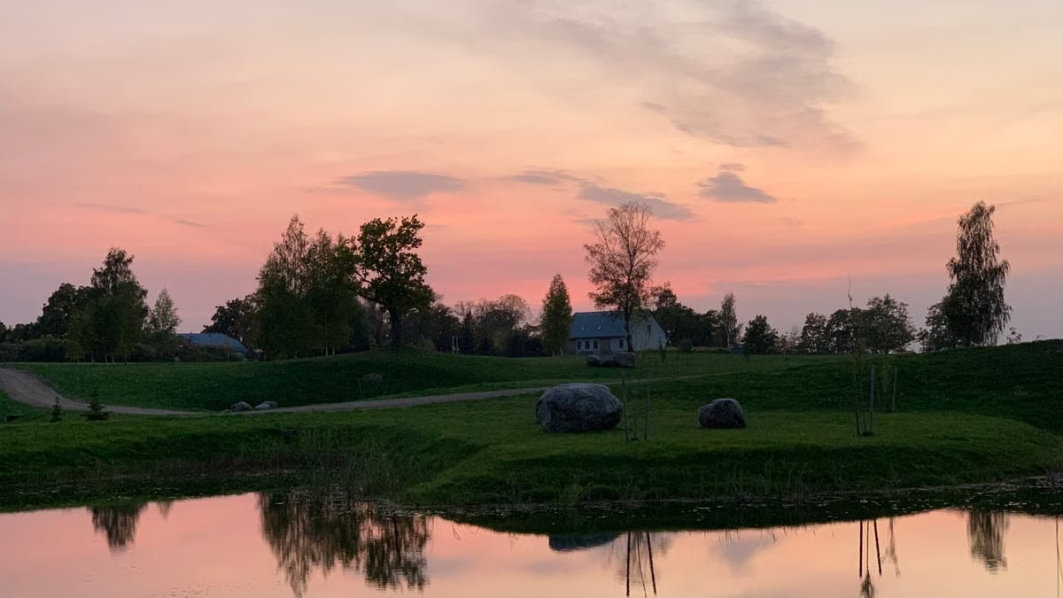 Scenic view of a serene sunset reflecting over a tranquil pond with silhouetted trees and distant house.